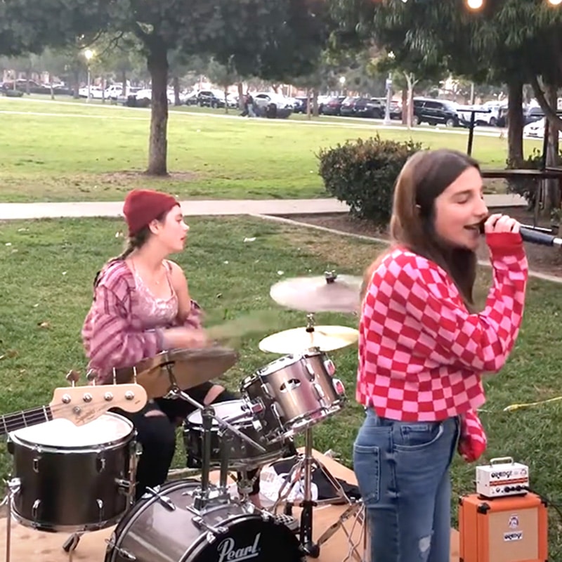 Two girls perform outdoors in a unique blend of music and creativity, embodying the spirit of Remix Education; one sings into a microphone wearing a red checkered sweater, while the other plays drums in a red beanie. They are set up on grass, with trees and parked cars in the background.