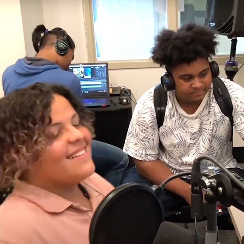 Three teens in a recording studio: one smiles at a microphone, another sits beside her, and the third works at a computer in the background wearing headphones, all united by their passion for music and its power to enhance literacy.
