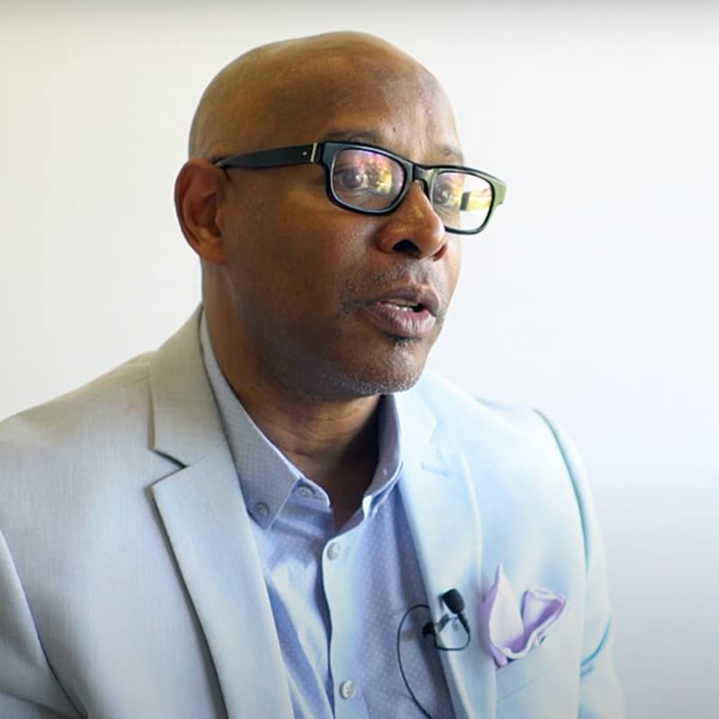 A man in glasses and a light blue suit jacket sits against a plain background, speaking with a microphone clipped to his shirt.