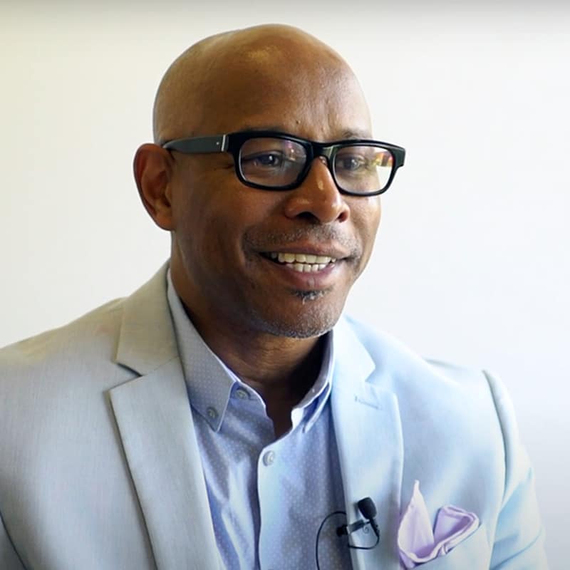 A man wearing glasses and a light blue suit jacket smiles while speaking, embodying the spirit of Learning Together. He has a lavender pocket square and a microphone clipped to his shirt. The background is plain and bright.