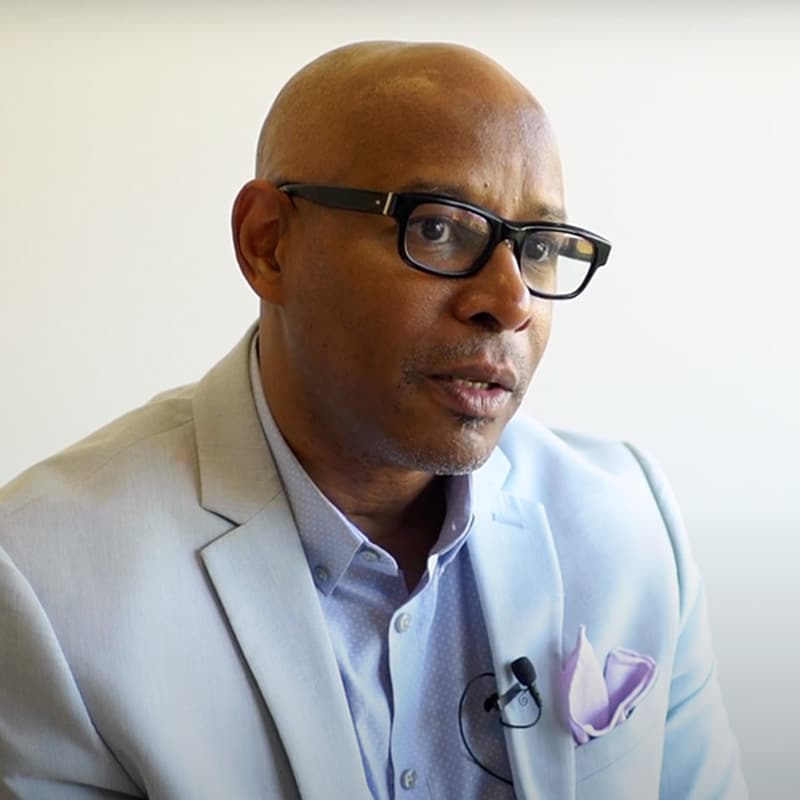 A man in a light blue suit jacket and glasses sits against a plain background, wearing a lavender pocket square and a microphone clipped to his lapel, ready to share his insights on contributing to the world.