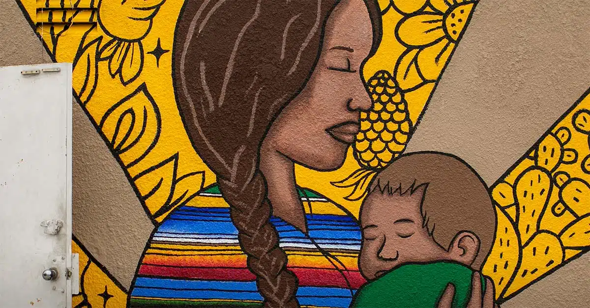 A colorful mural, featured in Issue 20 of Unboxed, shows a woman with a braid, eyes closed, holding a sleeping child. The background features yellow flowers, fruit, and geometric shapes.