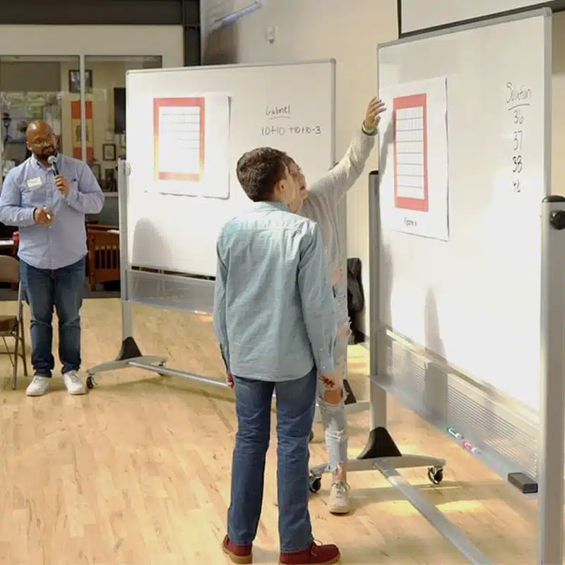 Three people are in a well-lit room with two large whiteboards. One person stands by a whiteboard explaining charts as part of a collaborative teaching session, while another listens attentively. A third person holds a microphone nearby, capturing insights into math education.