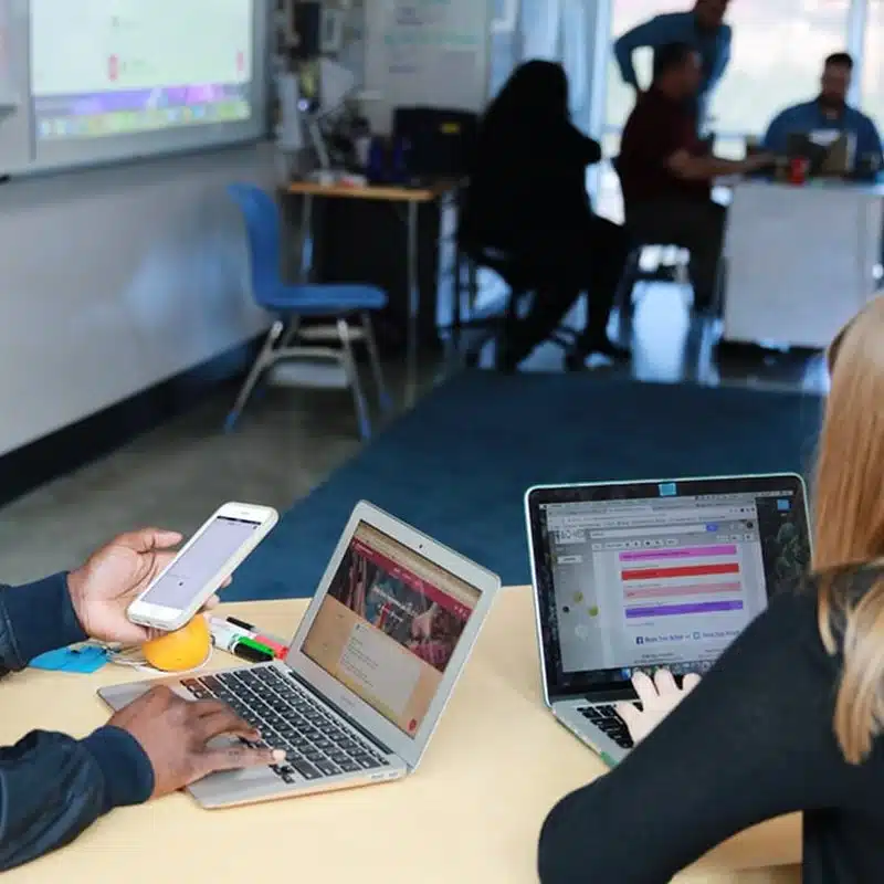 Two people use laptops at a classroom table, showcasing the integration of education technology in learning environments. One also holds a smartphone, emphasizing modern tools for both students and educators. In the background, several people sit together near a whiteboard and desks, some talking. A blue chair and classroom supplies are visible.