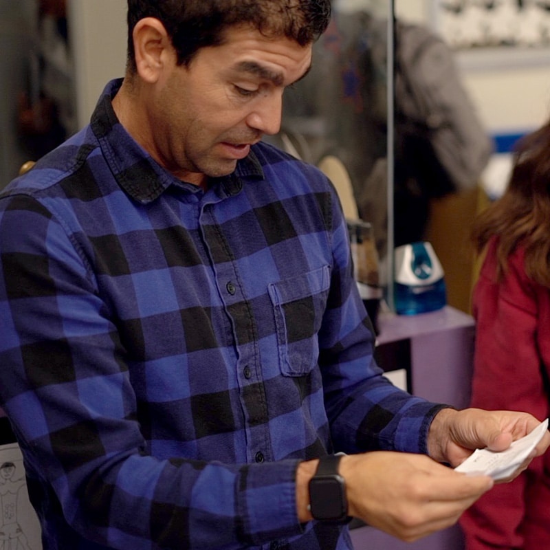 A man in a blue plaid shirt looks surprised while reading a piece of paper, possibly reflecting on recent assessment practices. He stands indoors, with shelves and another person blurred in the background.