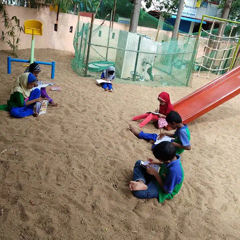 Five children sit on sandy ground in a playground, reading or writing in notebooks. Playground equipment, including a slide and climbing structure, is visible in the background—an ideal scene for little Bookworms to gather and enjoy their stories together.