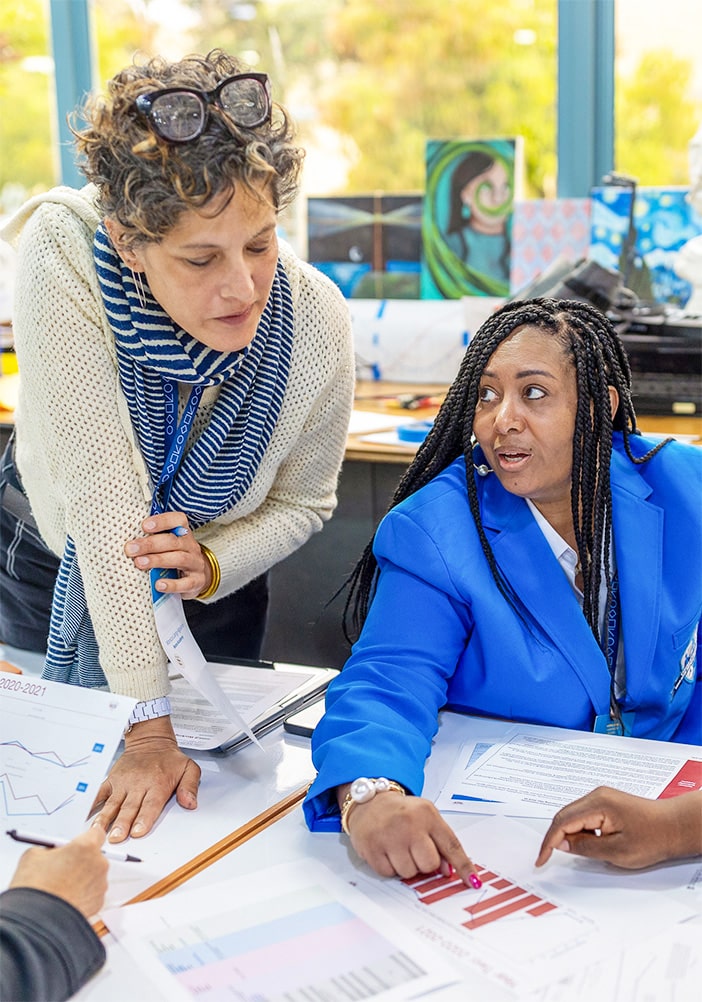 Two women talk at a table covered with charts and papers. One stands and listens while the other, in a blue blazer, points to a document—both focused on Improvement in Education during a spirited discussion in the brightly lit room.