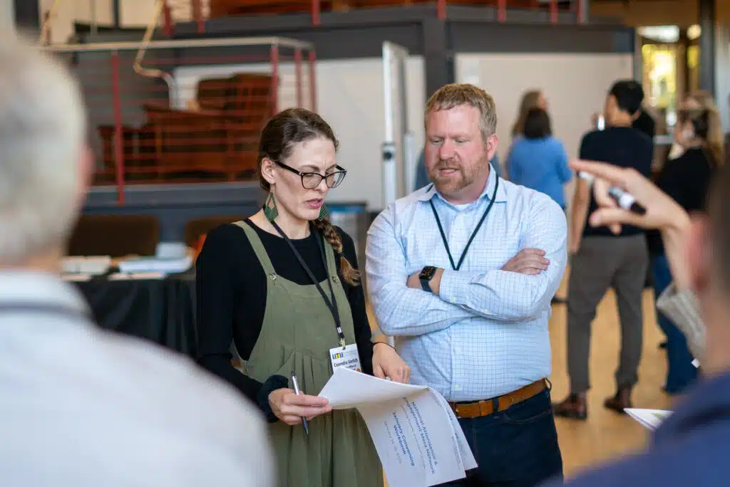 Two people are engaged in conversation at an indoor event. The woman, holding papers and sporting glasses and a green dress over a black top, discusses with the bearded man in a blue shirt about utilizing SEO strategies. Other attendees from the META Network are blurred in the background.