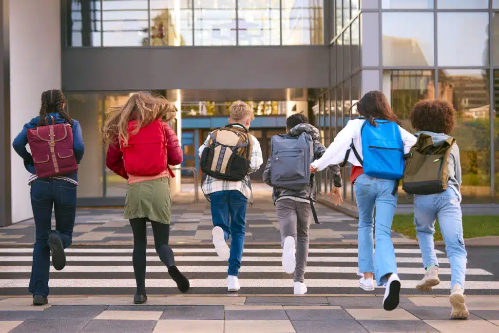 Six students with backpacks are running toward the entrance of a modern building, likely part of the RAISE Network. They cross a pedestrian area, their backs to the camera, in what appears to be an urban or campus setting on a sunny day.