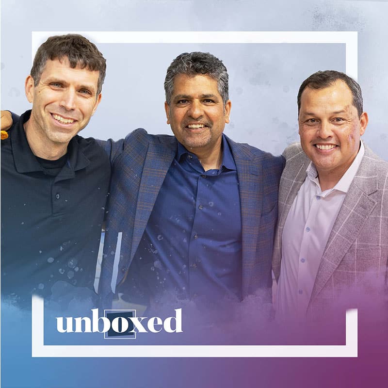 Three men, smiling and standing close together in business casual attire, pose for a photo against a light background. A white frame and the word unboxed, highlighting deeper learning, are overlaid at the bottom of the image.