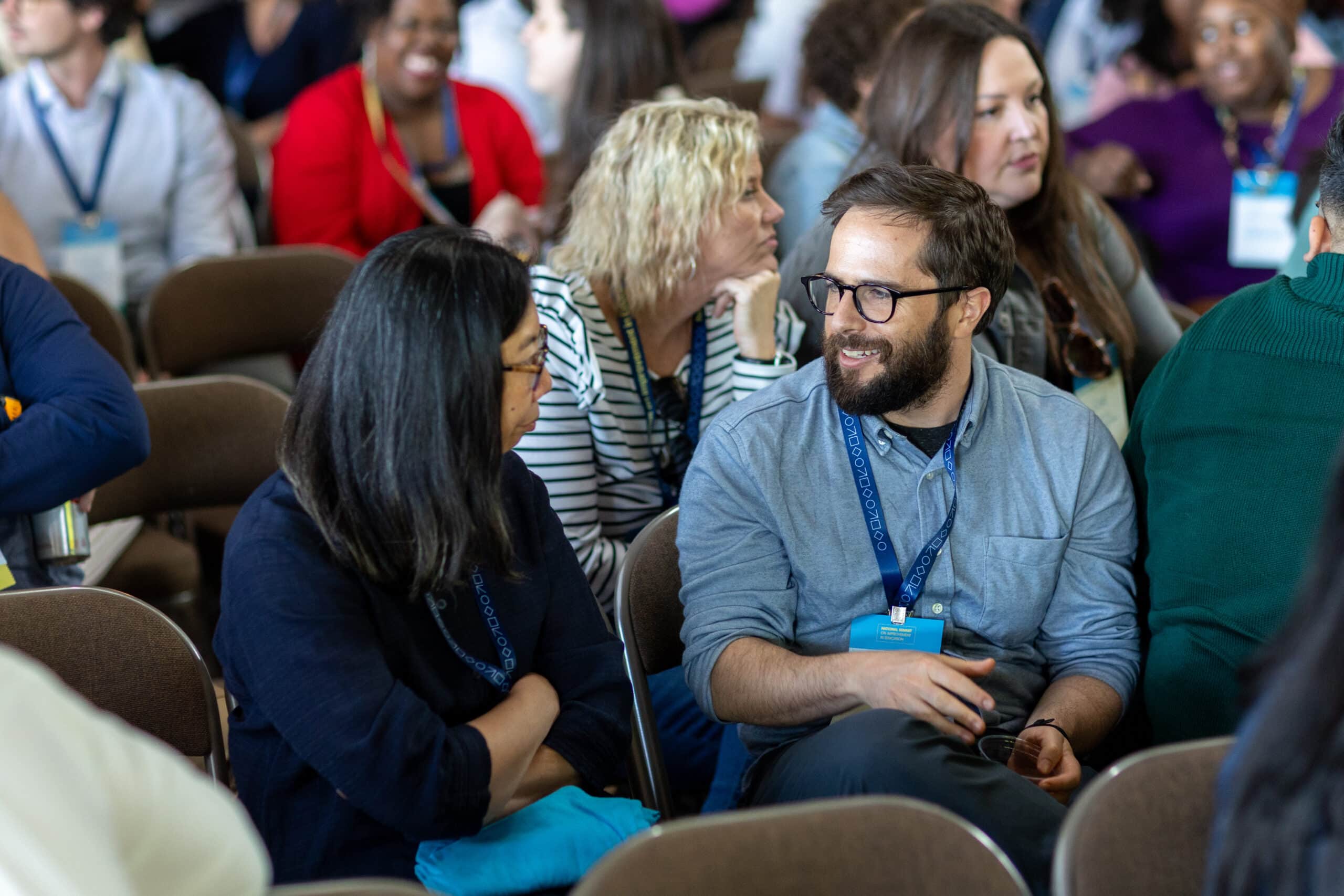 A group of people sit and talk in a conference room during the National Summit on Education. Two people, a man and a woman, are engaged in conversation in the foreground, while others with conference badges interact in the background.