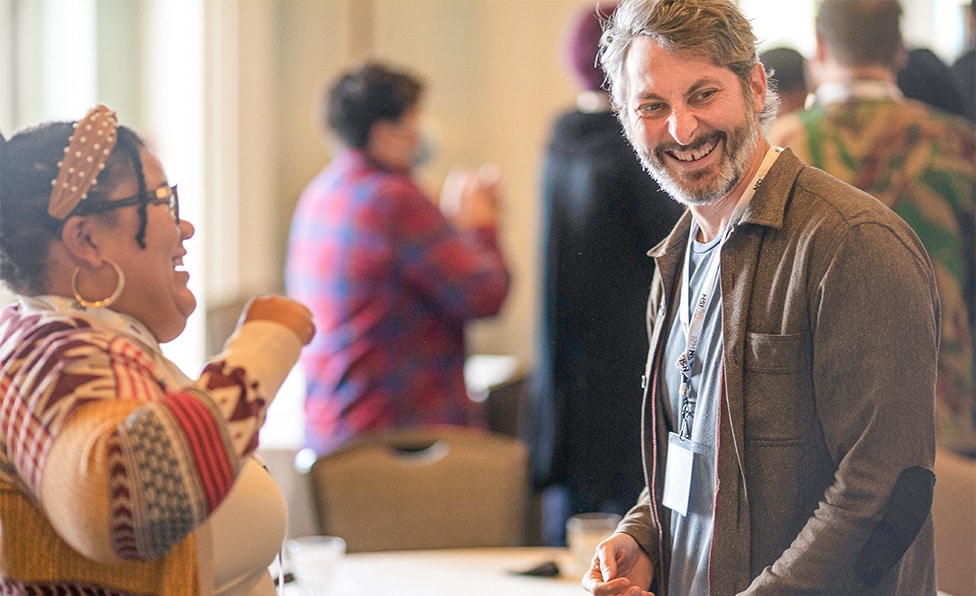 A man and a woman smiling and laughing together at an indoor event, with other people in the blurred background. The friendly, social atmosphere reflects the spirit of Kaizen and continuous improvement in their interactions.