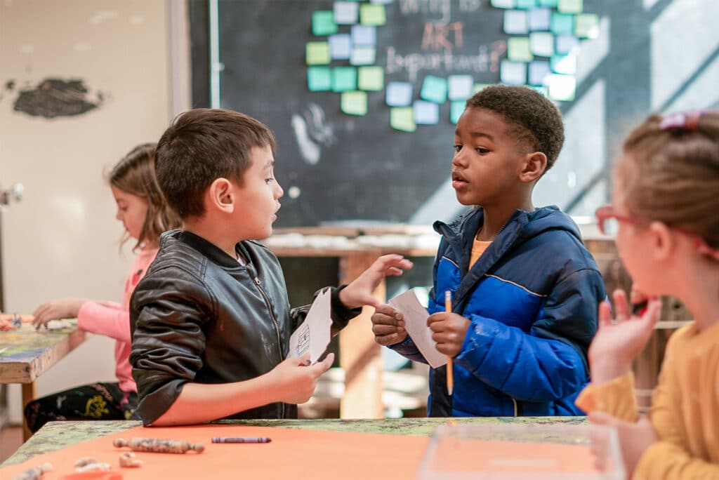 Four children are gathered around a table in a classroom, discussing priority areas and activities. Two boys in the center are talking and holding papers, while two girls sit nearby. A chalkboard with colorful notes is in the background.