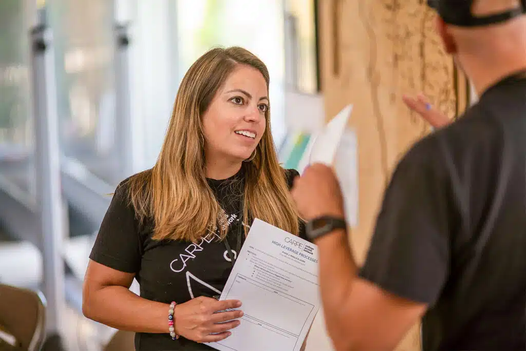 A woman holding papers smiles and talks to a person whose face is not visible. They appear to be discussing priority areas in an indoor setting filled with natural light.