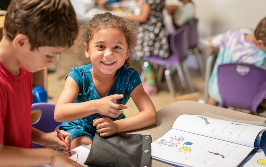 A young girl smiles at the camera while sitting at a classroom desk with a workbook open in front of her. Other children are seated nearby, engaged in activities. The bright, colorful setting invites you to Submit A Story about this joyful moment.
