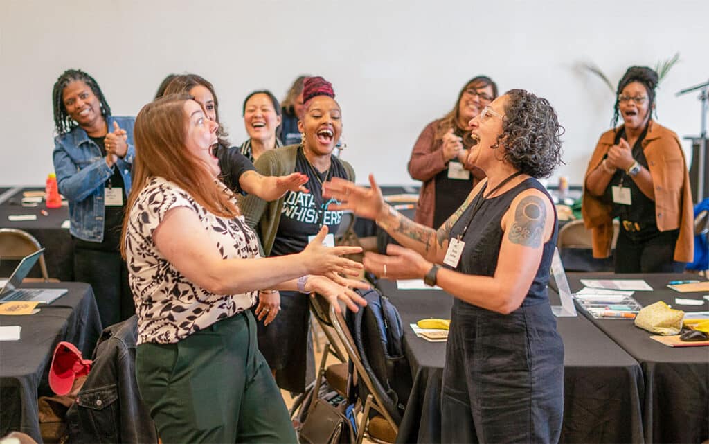 A group of women stand in a conference room, laughing and smiling enthusiastically as two women in front reach out toward each other, ready to share event details and complete the Event Form. Tables with papers and laptops are in the background.
