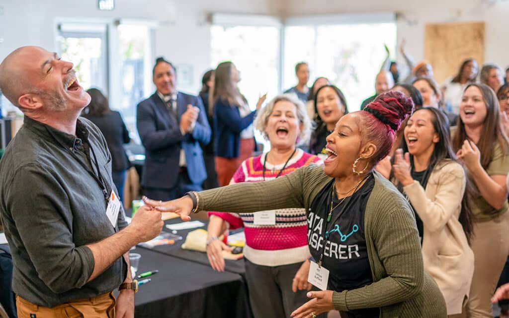 A group of people indoors enthusiastically laughing and cheering. Two people in the front are shaking hands and smiling, celebrating a successful Partnership Application, while others in the background clap at this lively workshop or event.