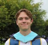 Nathan Ewart, a young person wearing a graduation gown and sash, stands outdoors in front of green foliage, smiling at the camera.