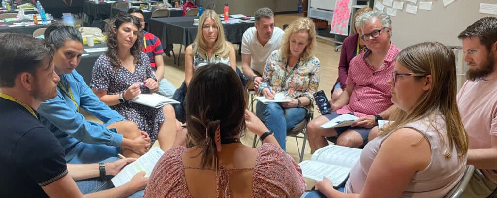 A group sits in a circle indoors, attentively listening and taking notes. The scene suggests an in-person collaborative discussion or workshop, likely part of the PBL Design Camp planned for May 2026.