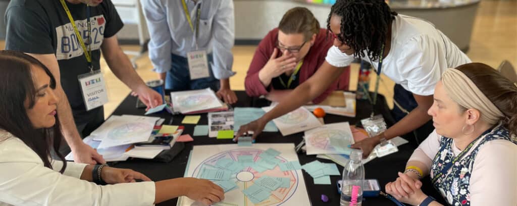 Six adults sit and stand around a table at the in-person PBL Design Camp, collaborating and discussing colorful circular charts and notes. Some are pointing at the charts, while others write or listen attentively during this Sept 2025 session.