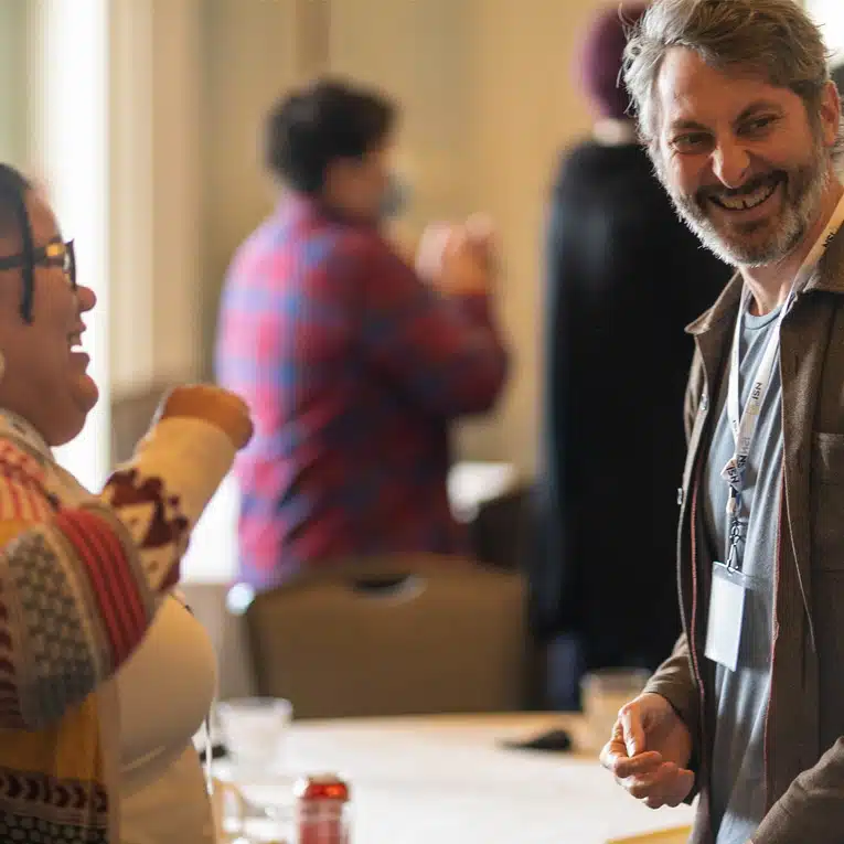 Two people stand and smile at each other in a brightly lit room during what appears to be a social or professional gathering. Other attendees are visible in the blurred background.