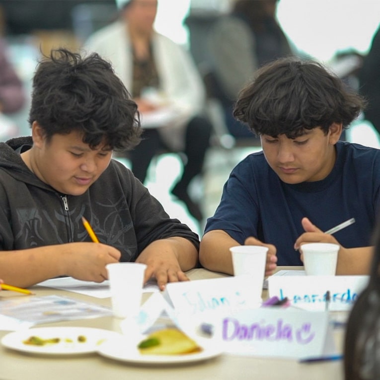 Two young people sit at a table, focused on writing or drawing with pencils. Papers, cups, and name tags, including one reading Daniela, are on the table. Other people are visible in the blurred background.