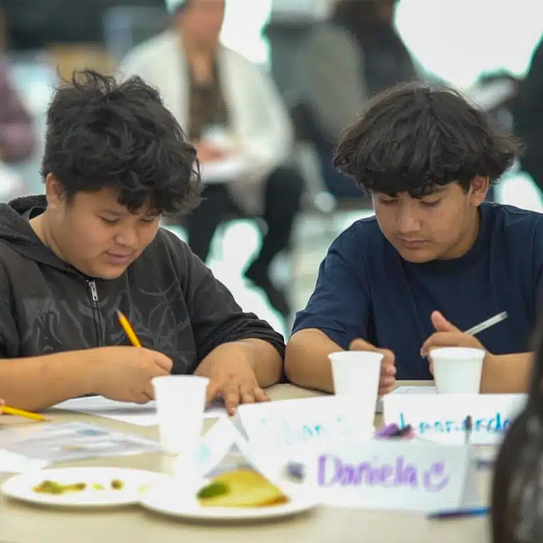 Two young people sit at a table, focused on writing or drawing with pencils. Papers, cups, and name tags, including one reading Daniela, are on the table. Other people are visible in the blurred background.