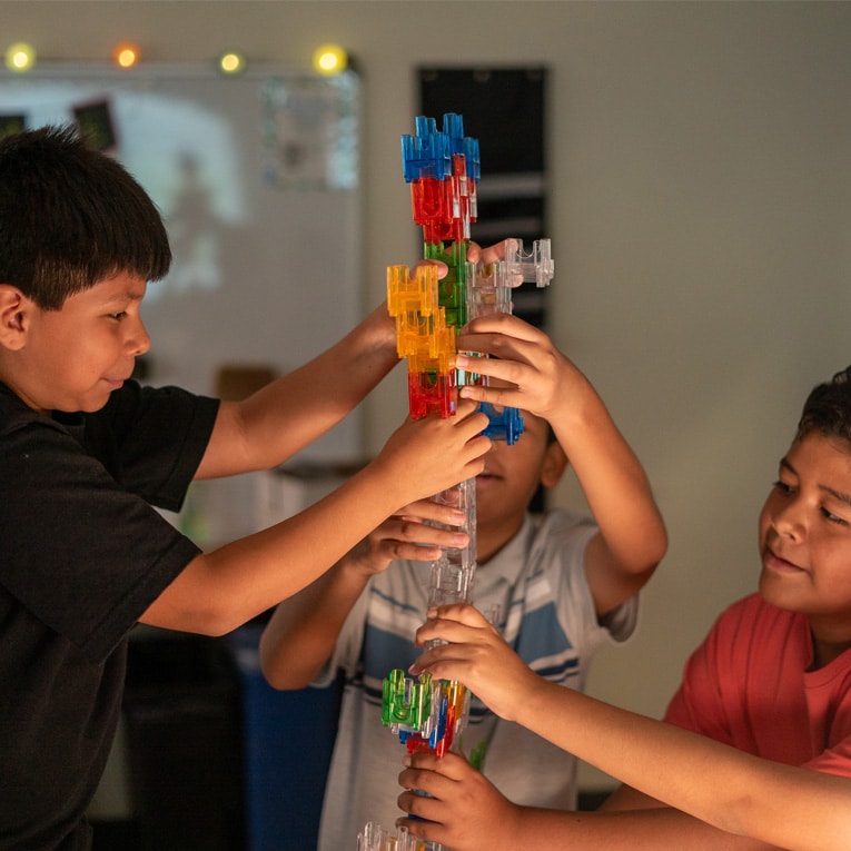 Three children work together to build a tall, colorful tower using translucent interlocking plastic pieces in a classroom setting.