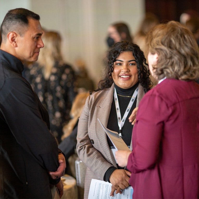 Three people stand in a group, smiling and talking at an indoor social or professional event. One woman holds papers, and all wear name badges on lanyards. Other attendees are visible in the blurred background.