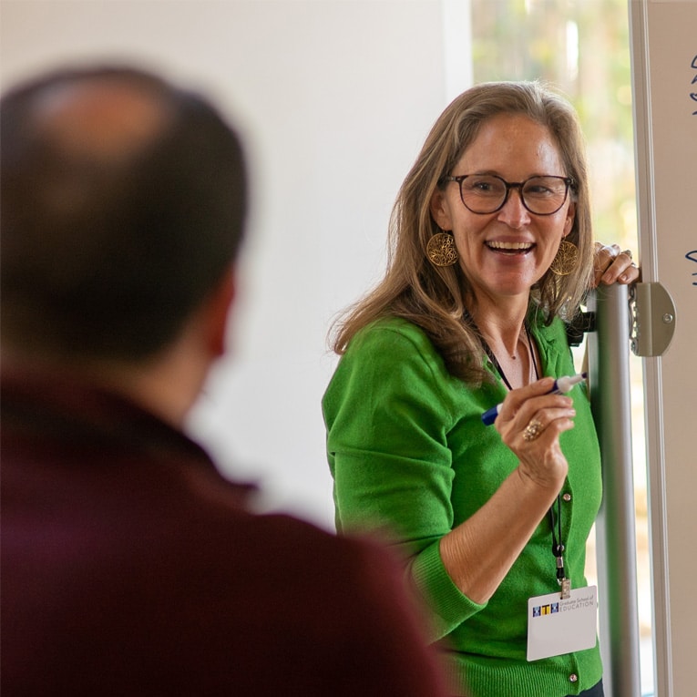 A woman in a green sweater, wearing glasses and large earrings, smiles and gestures with a marker while standing at a whiteboard, facing a person in the foreground.