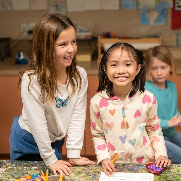 Two young girls smiling and standing at a table with pencils and paper, one wearing a white shirt with a butterfly, the other wearing a hoodie with colorful hearts. Another child sits in the background.