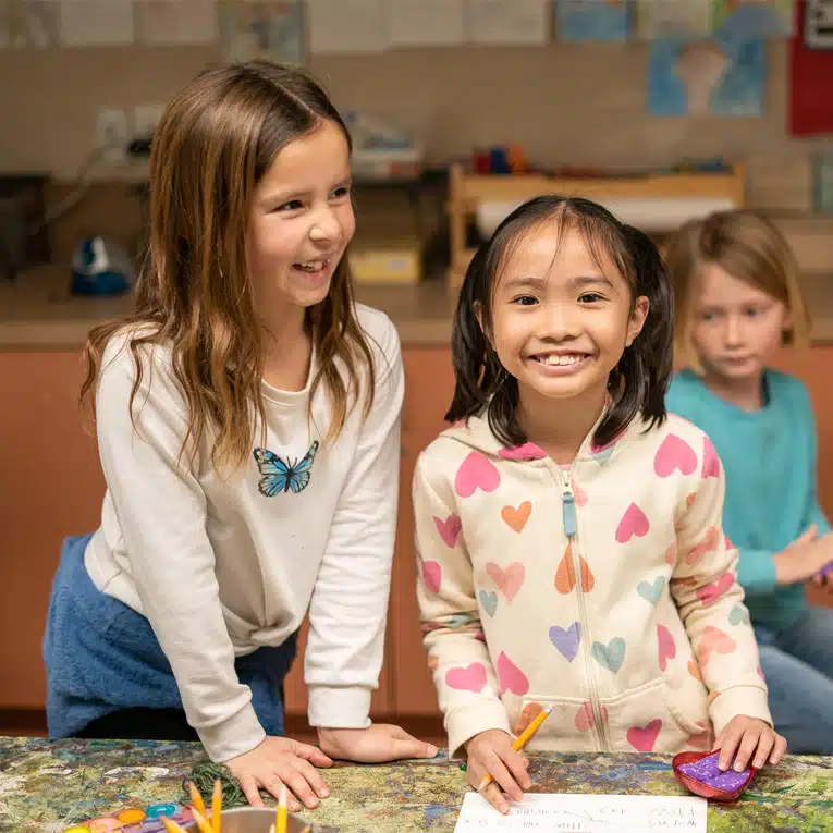Two young girls smiling and standing at a table with pencils and paper, one wearing a white shirt with a butterfly, the other wearing a hoodie with colorful hearts. Another child sits in the background.