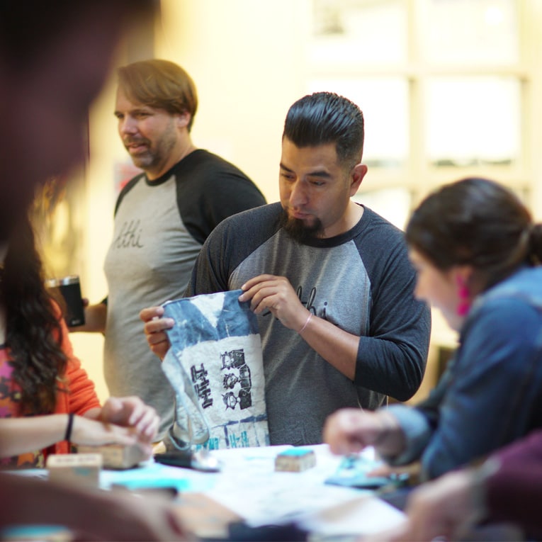 A man in a gray and black shirt holds up a printed cloth while others gather around a table, engaging in a creative group activity. The scene is indoors with warm lighting and a collaborative atmosphere.