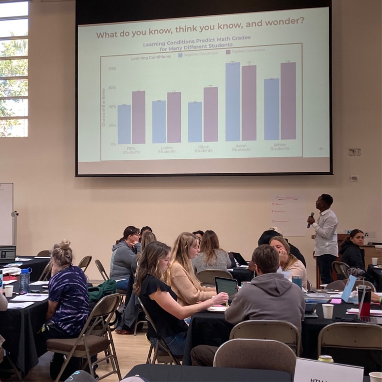 A presenter stands near a projected chart explaining learning conditions and math grades to a group of seated attendees in a classroom, with people discussing and taking notes at round tables.