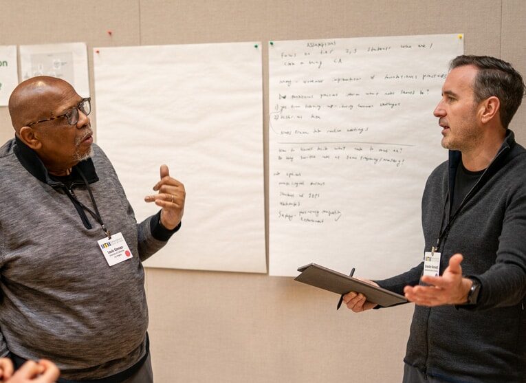 Two men wearing conference badges have a discussion in front of large sheets of paper with handwritten notes, possibly during a workshop or brainstorming session. One man gestures while the other holds a tablet.