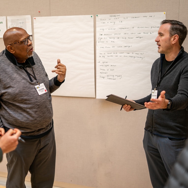 Two men wearing conference badges have a discussion in front of large sheets of paper with handwritten notes, possibly during a workshop or brainstorming session. One man gestures while the other holds a tablet.