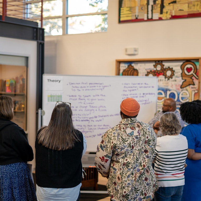 A group of people stand facing a whiteboard, listening to a presenter in a brightly lit room decorated with colorful art. The whiteboard is filled with handwritten notes.