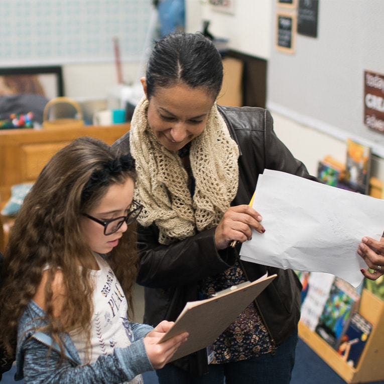 A woman and a young girl stand together in a classroom, looking at papers and smiling. The girl holds a clipboard and wears glasses, while the woman wears a scarf and jacket. Shelves and posters are visible in the background.