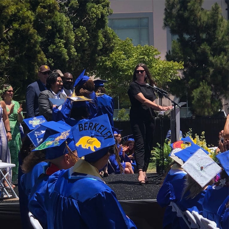 A woman in black speaks at an outdoor graduation ceremony, facing students in blue caps and gowns. Some caps are decorated, one with Berkeley and a bear. Trees and a building are in the background.