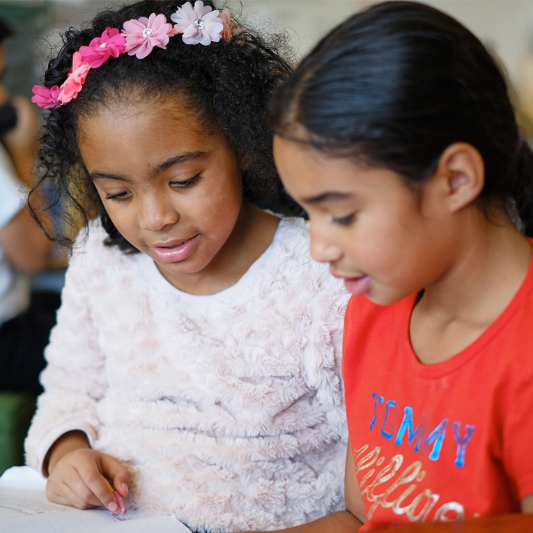 Two young girls sit closely together reading a book. One wears a pink flower headband and a fluffy white top, while the other wears a red shirt. They appear focused and engaged with the book.