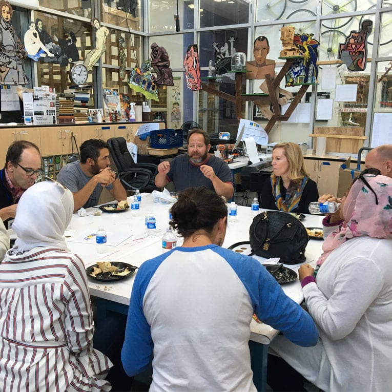 A group of people sit around a table sharing a meal and talking in a creative, art-filled workspace with colorful decorations and artwork on the walls. Bottled water and plates of food are on the table.