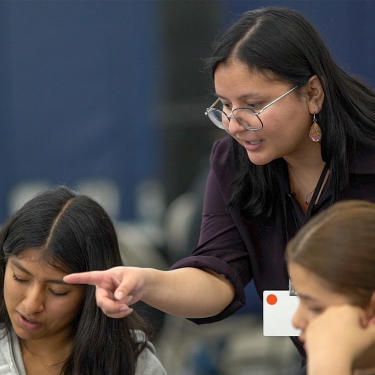 A woman with glasses leans over and points at something on a table, explaining to two young women who are looking down and listening attentively. The setting appears to be a classroom or workshop.