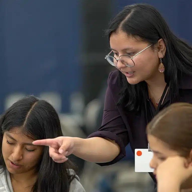 A woman with glasses leans over and points at something on a table, explaining to two young women who are looking down and listening attentively. The setting appears to be a classroom or workshop.