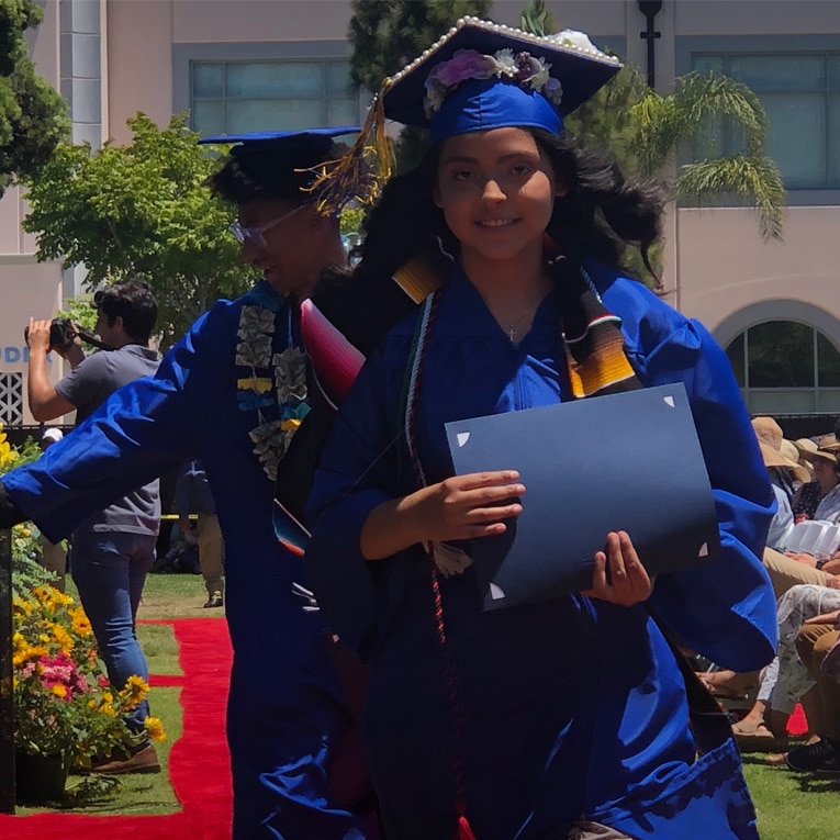 A student in a blue graduation gown and cap walks outdoors holding a diploma, smiling. Another graduate in similar attire is behind her. People are seated and watching the ceremony under a sunny sky.