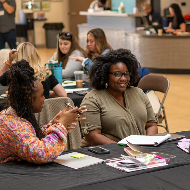 Two women sit at a table talking, with notebooks and phones in front of them. Other people are seated and working in the background in a bright, casual indoor setting.