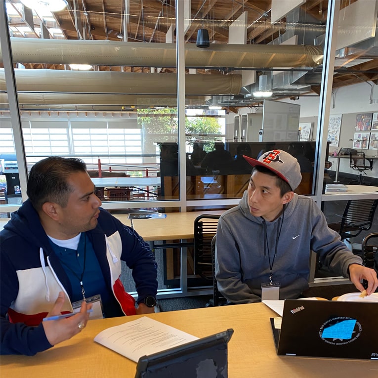Two men sit at a table in a modern office, engaged in discussion with papers and laptops in front of them. One man gestures with a pen while the other listens attentively. Large windows and high ceilings are visible in the background.
