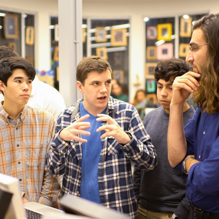 Four young men stand together indoors, engaged in a lively discussion. One is speaking animatedly with his hands, while the others listen attentively. Framed pictures are visible in the background.