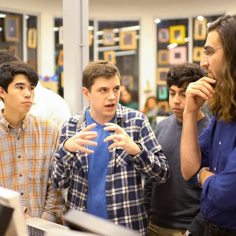 Four young men stand together indoors, engaged in a lively discussion. One is speaking animatedly with his hands, while the others listen attentively. Framed pictures are visible in the background.
