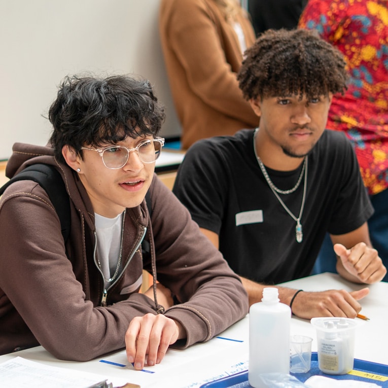 Two young men sit at a table in a classroom setting, appearing engaged in a discussion or activity. Papers, a pen, and plastic containers are on the table. Other people are visible in the background.