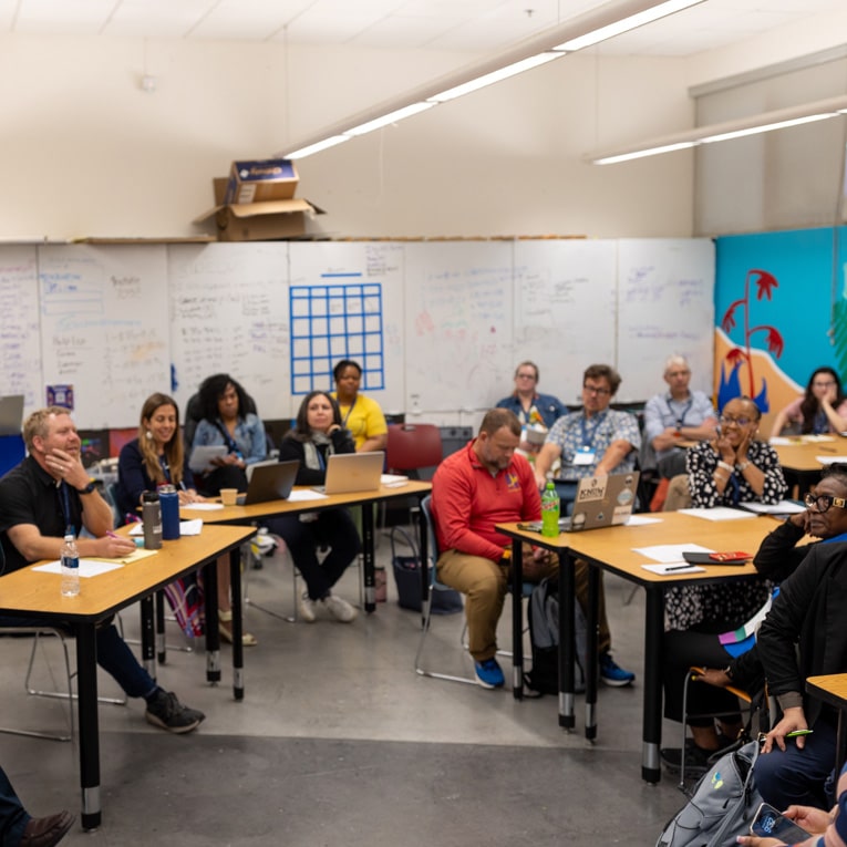 A group of adults sitting at tables in a classroom, listening attentively to a speaker out of frame. Laptops, notebooks, and water bottles are on the tables. Whiteboards with notes fill the walls.